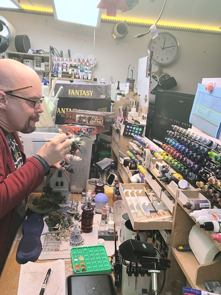 Baldie in his workshop between piles of boxes with mini's and racks full of paint.